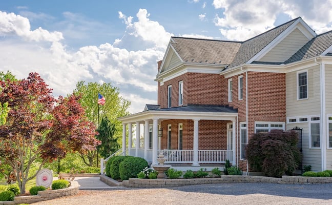 Red brick colonial house with white columns, front porch, and attached garage, surrounded by landscaped gardens with flowering trees and shrubs.