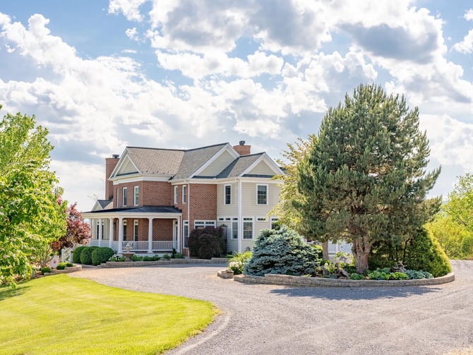 Two-story brick and cream colonial house with front porch, circular driveway, manicured landscaping, and tall evergreen trees.